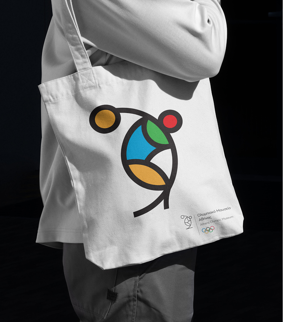 A person carrying a white canvas tote bag featuring a colorful geometric illustration of a discus thrower and the Athens Olympic Museum logo.